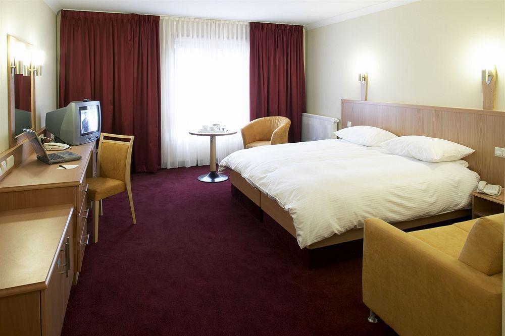 Clayton Hotel Dublin Airport Coolock Phòng bức ảnh The photo shows a hotel room featuring a double bed with white linens and pillows. There are two bedside lamps mounted on the wall above the bed. To one side, there's a small round table with two chairs. Opposite the bed, a wooden desk with a chair i